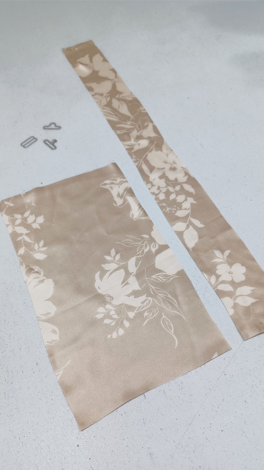 A long strip of floral silk fabric and a smaller rectangular piece alongside silver bow tie hardware on a work table.