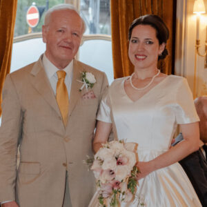 A close-up thumbnail portrait of the bride in her short-sleeved ivory silk Duchess satin wedding gown with a v-neck, next to the groom in his beige suit with a yellow tie, holding a rose and lily bouquet in a London reception room.