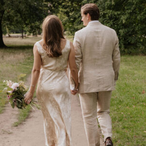 A bride and groom walking hand-in-hand in a park; the bride wears a floral silk dress tailored to fit her silhouette.