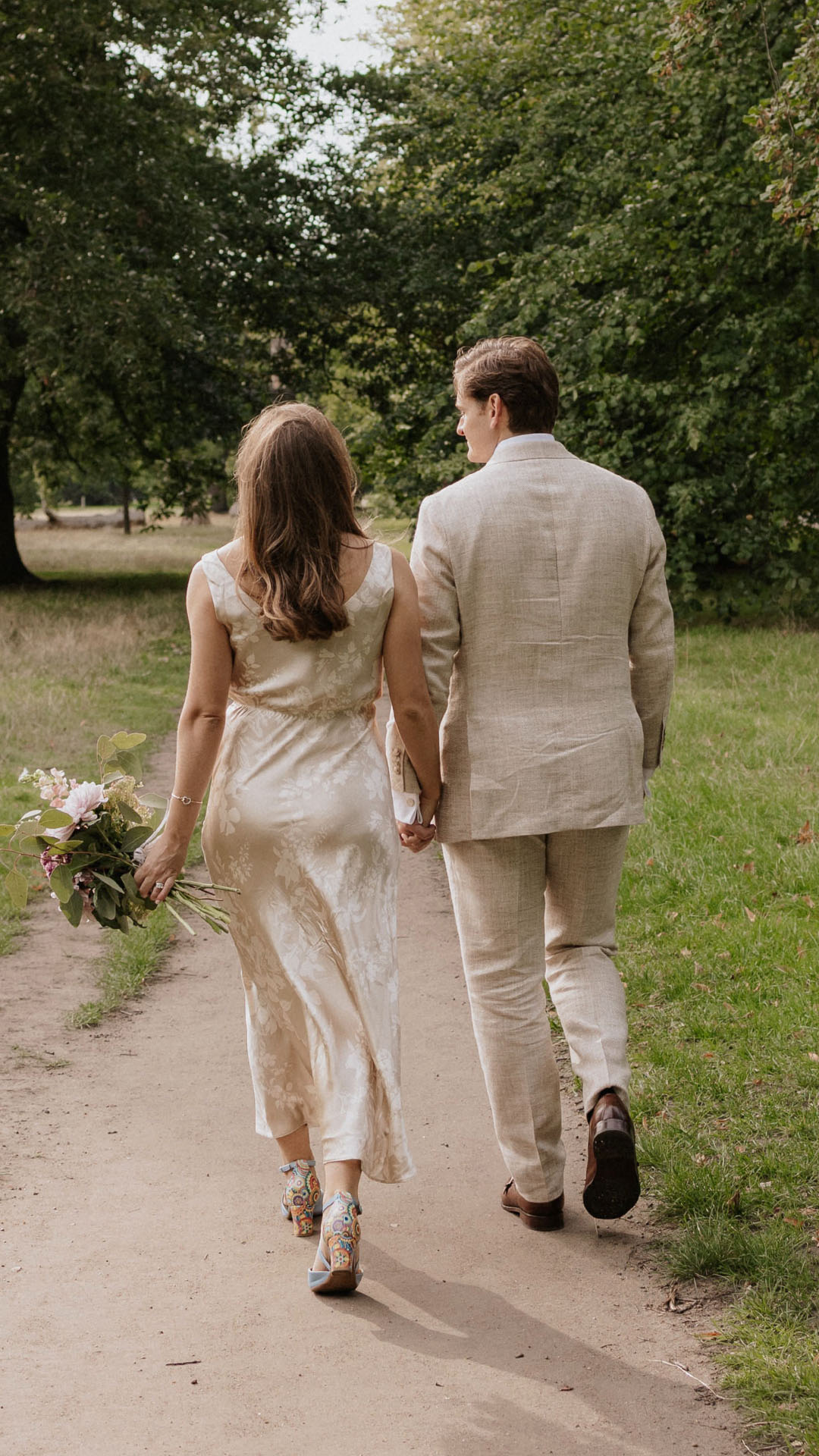 A bride and groom walking hand-in-hand in a park; the bride wears a floral silk dress tailored to fit her silhouette.